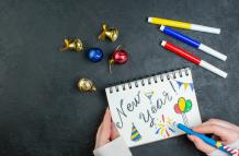 top-view-of-hand-holding-pen-on-spiral-notebook-with-new-year-writing-and-drawings-decoration-accessories-on-black-background