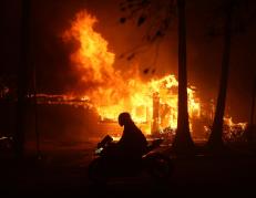 Una persona conduce una moto junto a una casa en llamas debido al incendio forestal de Palisades.