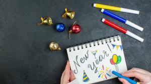 top-view-of-hand-holding-pen-on-spiral-notebook-with-new-year-writing-and-drawings-decoration-accessories-on-black-background