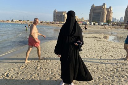 Una mujer árabe se pasea en la playa con su abaya y la burka cubriéndole todo el rostro, ante la curiosa mirada de un turista.