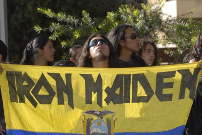 Un grupo de seguidores con cabelleras largas y gafas de sol, sosteniendo una enorme bandera amarilla con el logo de Iron Maiden y el escudo de Ecuador.