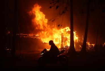 Una persona conduce una moto junto a una casa en llamas debido al incendio forestal de Palisades.