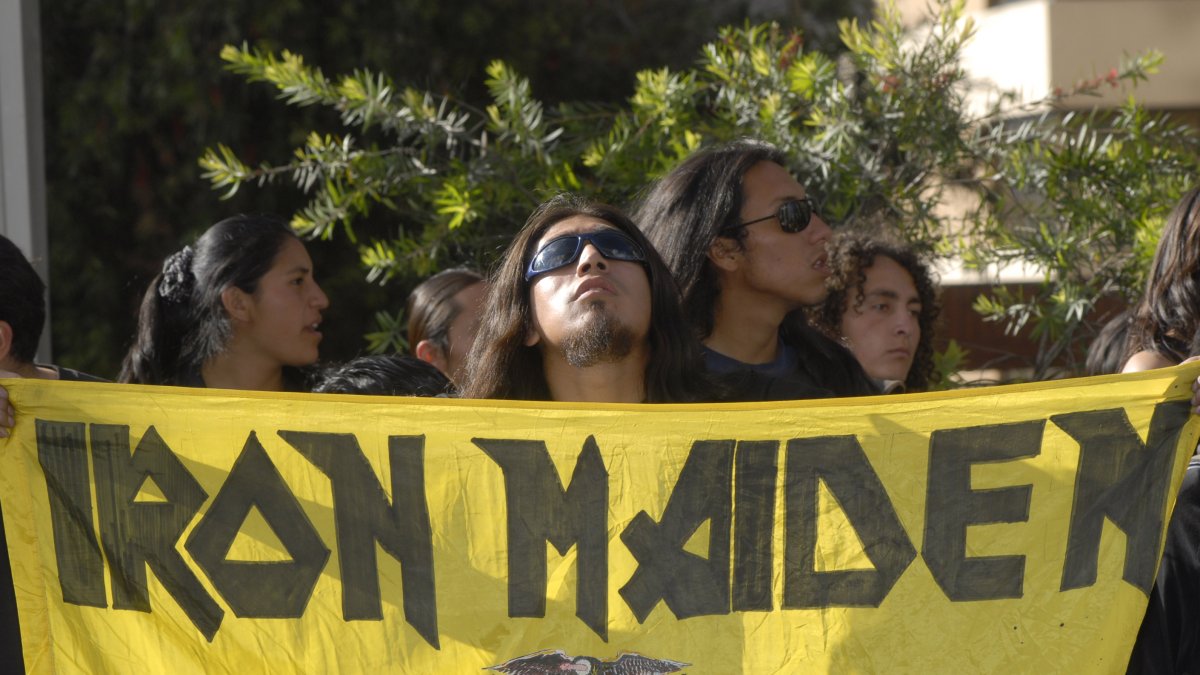 Un grupo de seguidores con cabelleras largas y gafas de sol, sosteniendo una enorme bandera amarilla con el logo de Iron Maiden y el escudo de Ecuador.