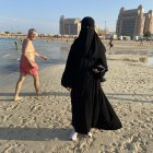 Una mujer árabe se pasea en la playa con su abaya y la burka cubriéndole todo el rostro, ante la curiosa mirada de un turista.